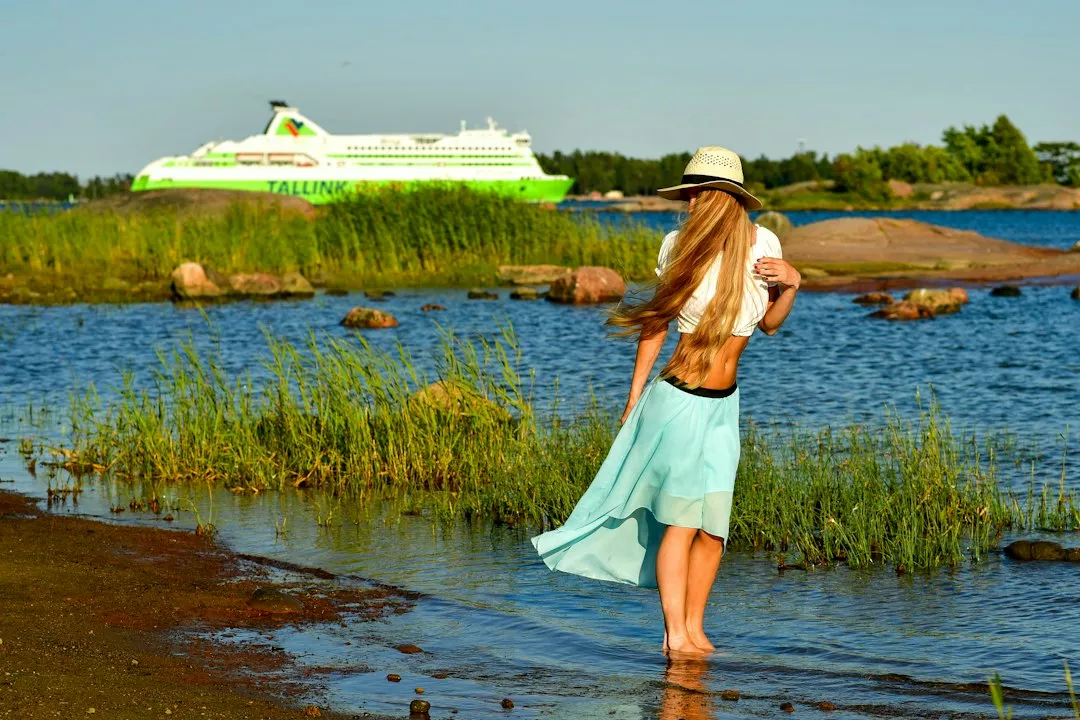 Woman in hat and skirt by water with ferry