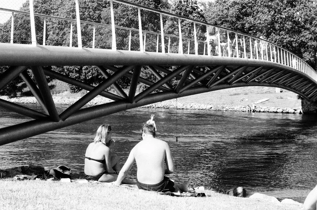 a man and a woman sitting on the bank of a river under a bridge
